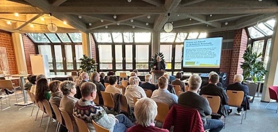 Bei der Veranstaltung „Wohnen mit Zukunft“ im Kreishaus SÜW ging es unter anderem um gemeinschaftliche Wohnformen im Alter. Foto: KV SÜW Menschen sitzen im Erdgeschoss der Kreisverwaltung und blicken auf eine Präsentationsfolie.