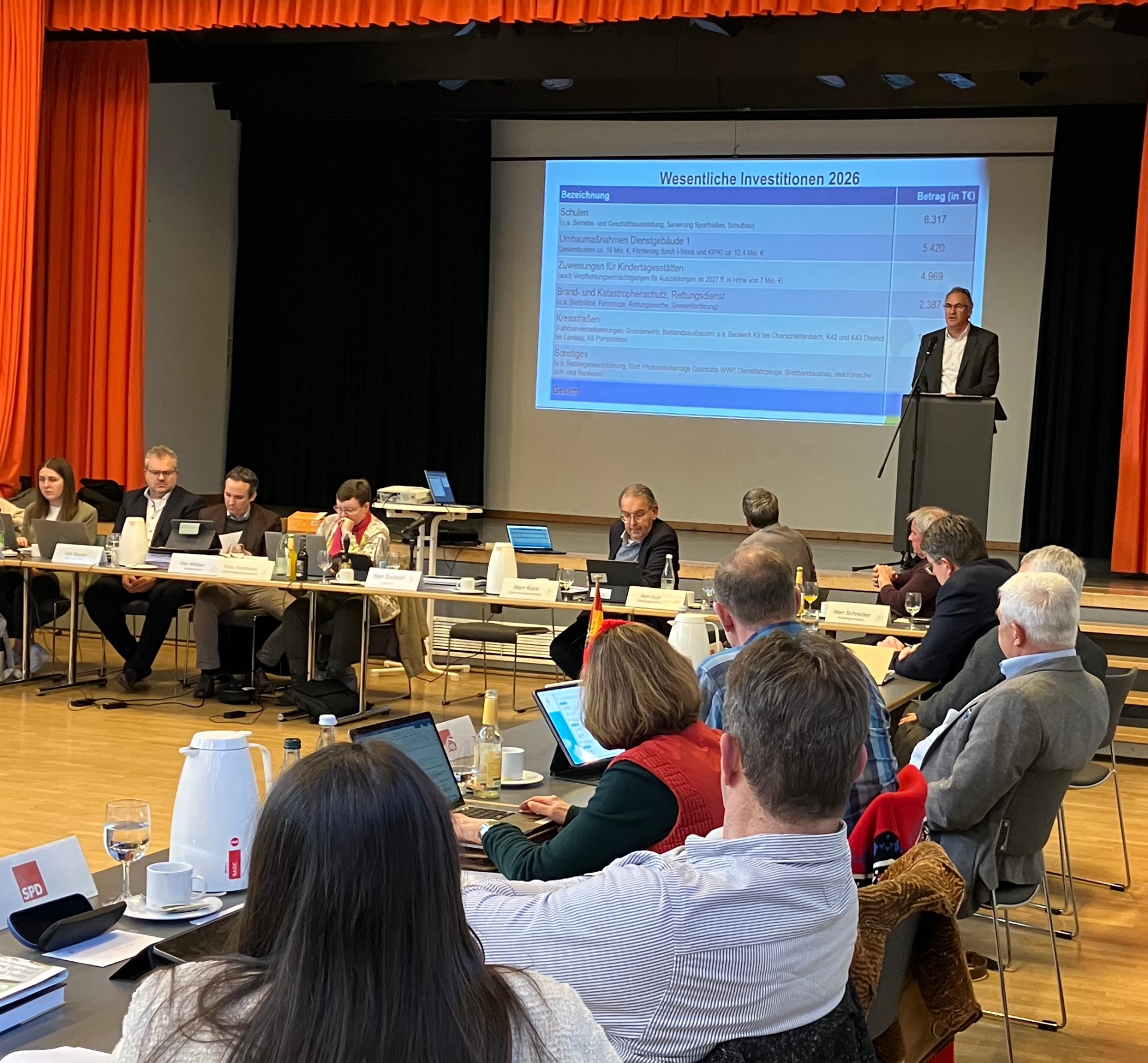 Blick in die bestuhle Mehrzweckhalle Hayna, Personen sitzen an Tischen, Dietmar Seefeldt hält am Pult eine Rede. Eine Präsentationsfolie zeigt die Überschrift "Wesentliche Investitionen".