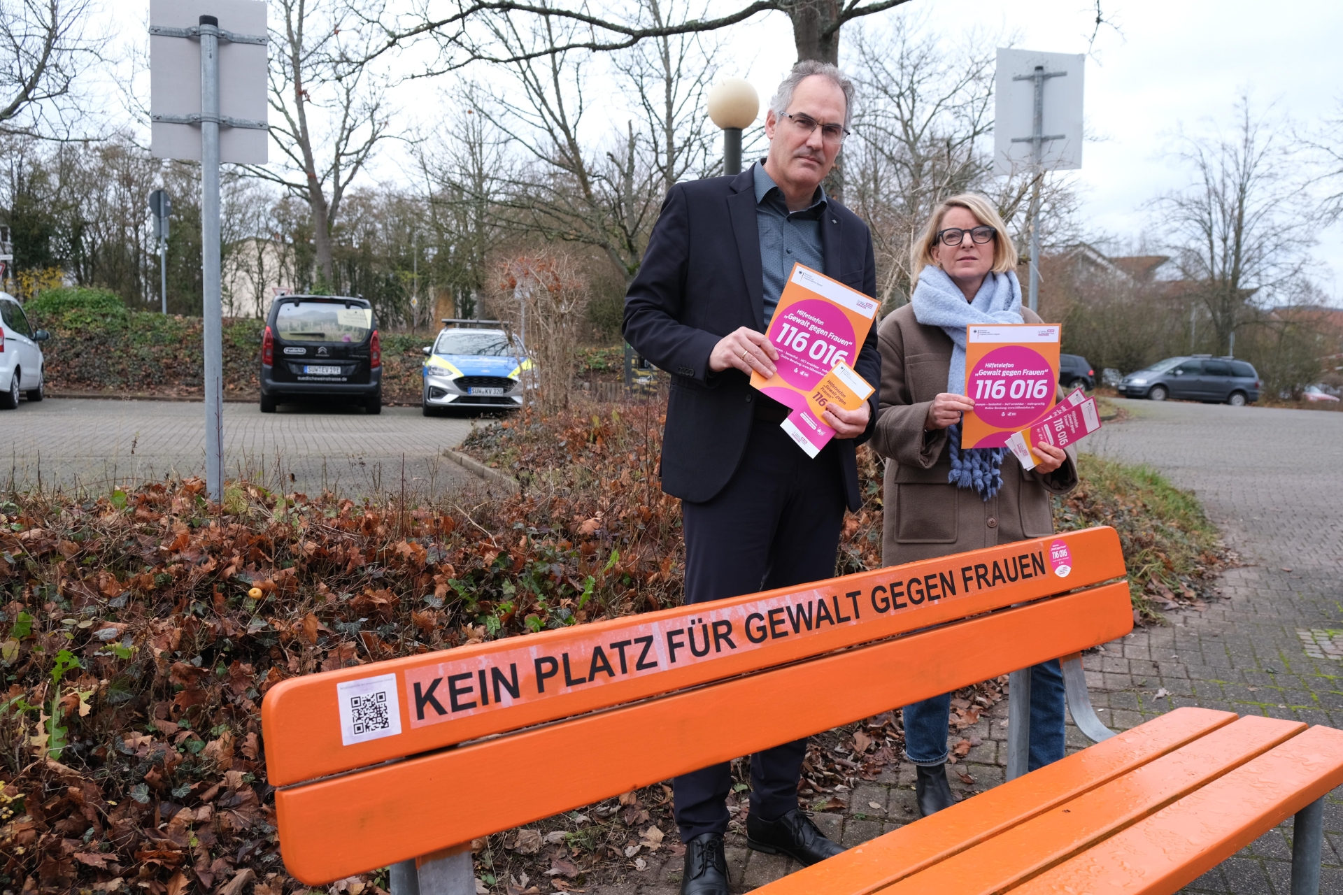 Landrat Dietmar Seefeldt und SÜW-Gleichstellungsbeauftragte Isabelle Stähle stehen mit ernsten Mienen hinter einer orangefarbenen Bank, auf der "Kein Platz für Gewalt gegen Frauen" steht, und halten Flyer zum Hilfetelefon in den Händen. Foto: KV SÜW