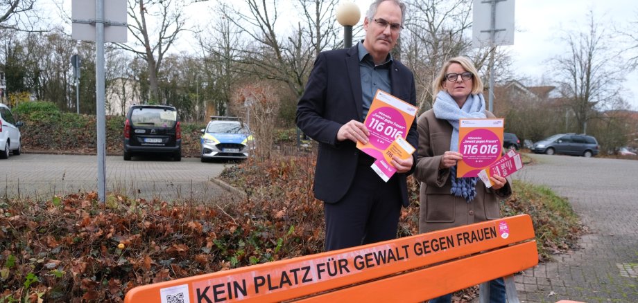 Landrat Dietmar Seefeldt und SÜW-Gleichstellungsbeauftragte Isabelle Stähle werben dafür, dass von Gewalt betroffene Frauen sich auch über die Feiertage rund um die Uhr an das Hilfetelefon wenden können. Foto: KV SÜW Landrat Dietmar Seefeldt und SÜW-Gleichstellungsbeauftragte Isabelle Stähle stehen mit ernsten Mienen hinter einer orangefarbenen Bank, auf der "Kein Platz für Gewalt gegen Frauen" steht, und halten Flyer zum Hilfetelefon in den Händen. Foto: KV SÜW