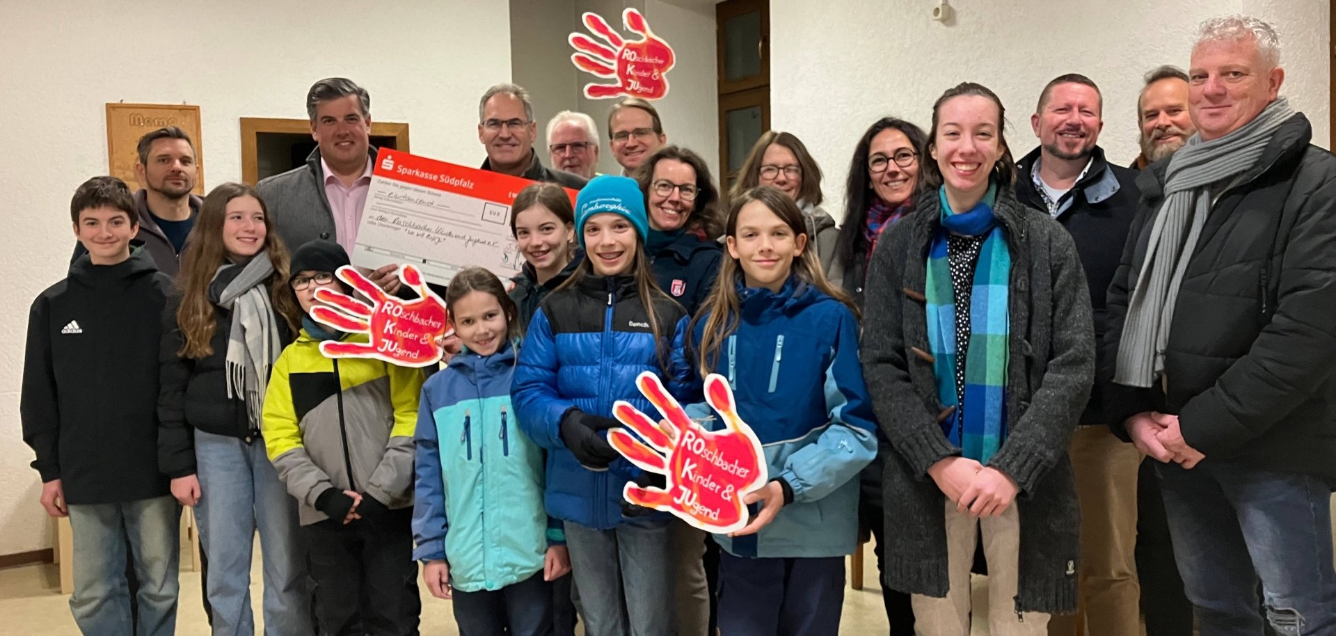 Gruppenbild mit zahlreichen Erwachsenen, Kindern und Jugendlichen. Einige Kinder halten eine große rote Hände aus Papier, die mit der Aufschrift "Roschbacher Kinder & Jugend" versehen sind. Eine Person hält einen großen symbolischen Scheck.