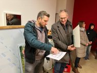 Architekt Kristoph Rheinwalt hält einen Bauplan in der Hand und zeigt darauf. Er erläutert offensichtlich den Plan für den nebenstehenden Landrat Dietmar Seefeldt, der den Plan betrachtet. Im Hintergrund sind eine Frau und ein Jugendlicher zu sehen.