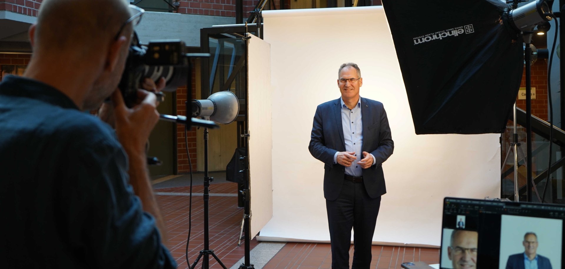 Landrat Dietmar Seefeldt steht im Foyer des Kreishauses vor einem weißen Fotohintergrund. Im Vordergrund sind sowohl technische Ausstattung des Fotoshootings zu sehen - Blitz, Softbox, Reflektor und ein Laptop, der auf einem Beistelltisch steht - als auch der Fotograf Thomas Brenner, der gerade das Porträt des Landrats ablichtet.