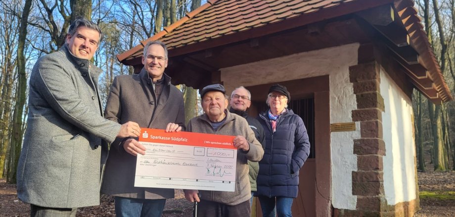 Fünf Personen stehen nebeneinander vor einem Bilderstock in einem Wald und halten gemeinsam einen großen symbolischen Scheck über 1.000 Euro. Alle blicken in die Kamera und lächeln.