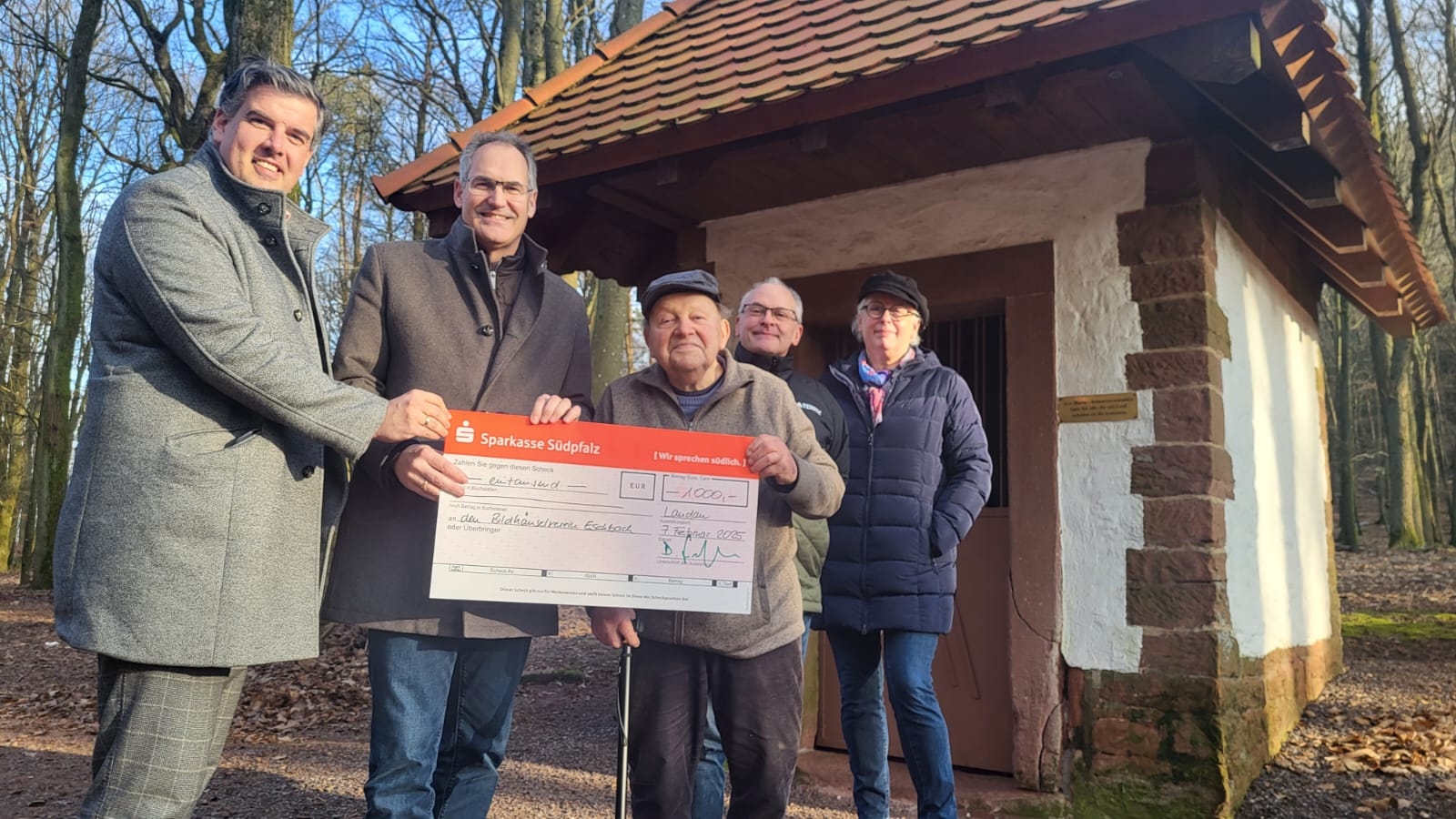 Fünf Personen stehen nebeneinander vor einem Bilderstock in einem Wald und halten gemeinsam einen großen symbolischen Scheck über 1.000 Euro. Alle blicken in die Kamera und lächeln.