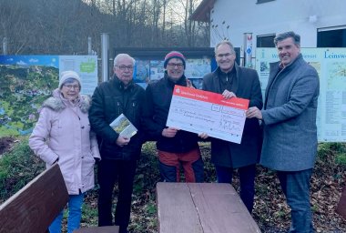 Fünf Personen stehen nebeneinander und halten gemeinsam einen großen symbolischen Scheck über 1.000 Euro. Alle blicken in die Kamera und lächeln.