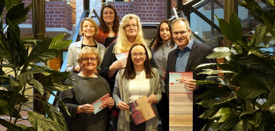 Marita Rothmann, Isabelle Stähle, Stephanie Heikamp, Inge Keller, Sandra Hubert, Nicole Kruppenbacher und Dietmar Seefeldt stehen auf der Treppe im Foyer des Kreishauses Südliche Weinstraße. Alle halten einen Flyer in der Hand, der das Programm der Veranstaltungsreihe "Brot und Rosen" 2025 enthält. Im Vordergrund sind links und rechts grüne Zimmerpflanzen zu sehen, die den Treppenaufgang flankieren.