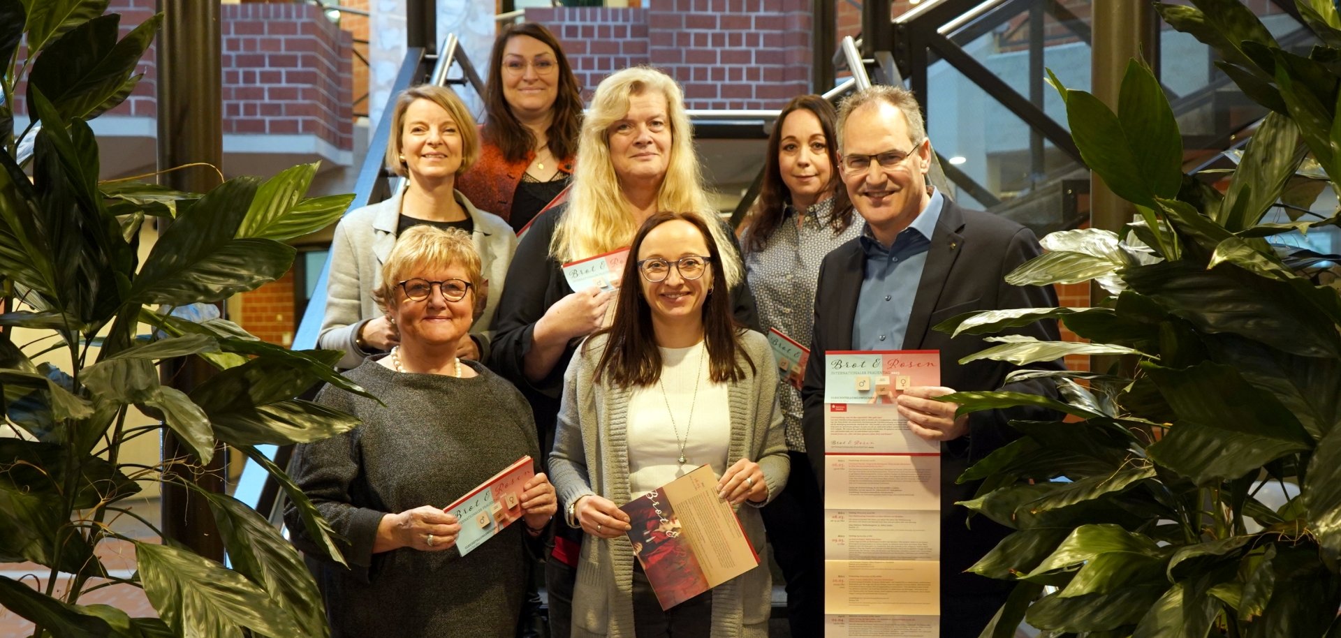Marita Rothmann, Isabelle Stähle, Stephanie Heikamp, Inge Keller, Sandra Hubert, Nicole Kruppenbacher und Dietmar Seefeldt stehen auf der Treppe im Foyer des Kreishauses Südliche Weinstraße. Alle halten einen Flyer in der Hand, der das Programm der Veranstaltungsreihe "Brot und Rosen" 2025 enthält. Im Vordergrund sind links und rechts grüne Zimmerpflanzen zu sehen, die den Treppenaufgang flankieren.