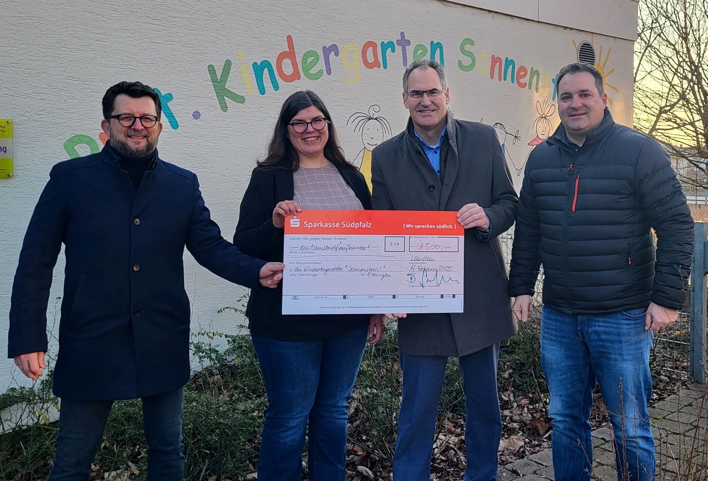 Vier Menschen vor einer Wand mit bunter Schrift, in der Hand einen übergroßen roten Spendenscheck.