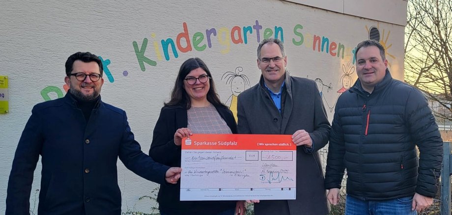 Vier Menschen vor einer Wand mit bunter Schrift, in der Hand einen übergroßen roten Spendenscheck.