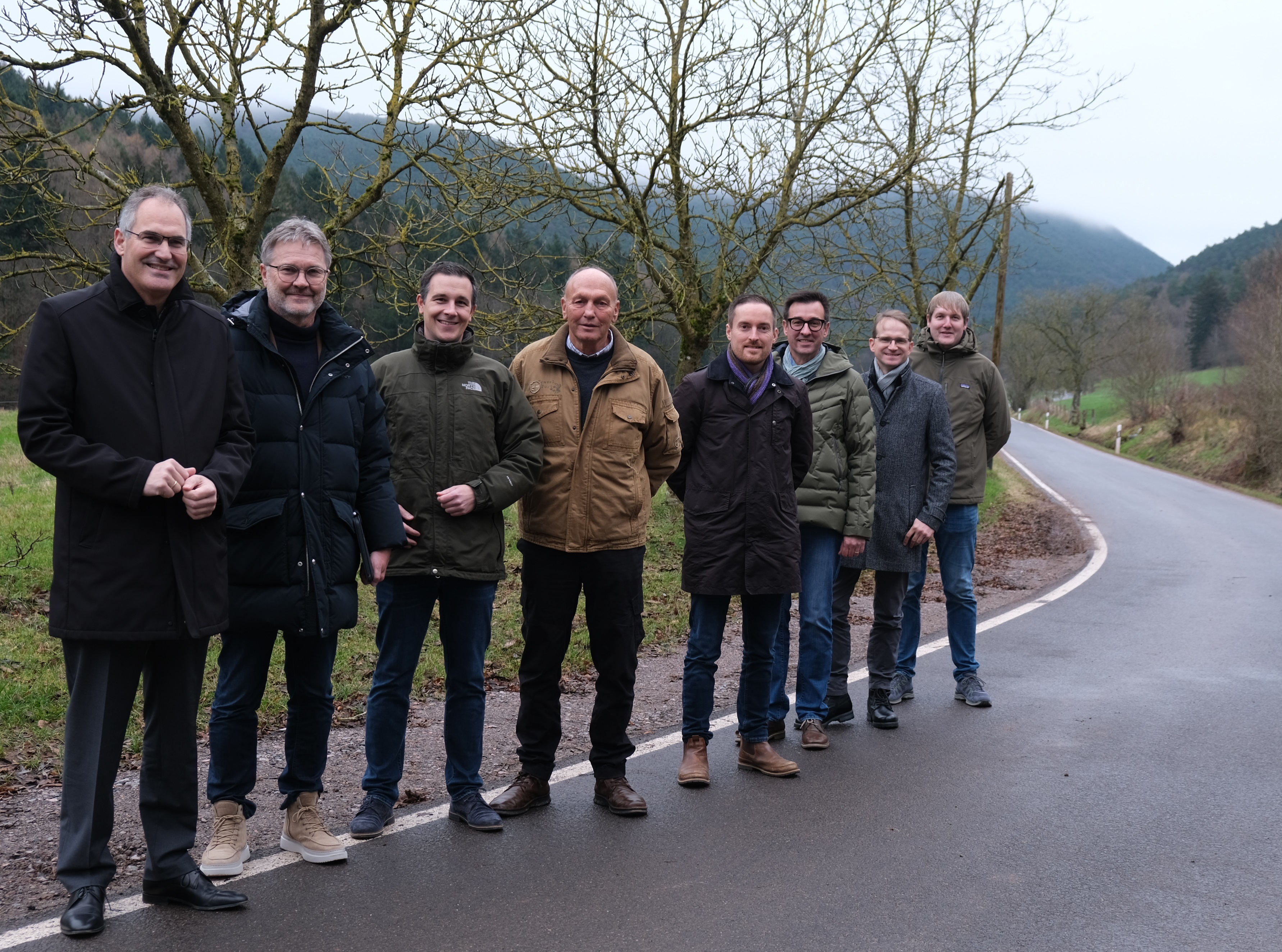 Männer stehen auf einer Straße, vor einer Kurve. Im Hintergrund Bäume und Hügel.