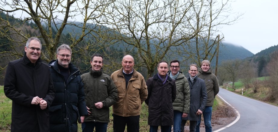 Männer stehen auf einer Straße, vor einer Kurve. Im Hintergrund Bäume und Hügel.