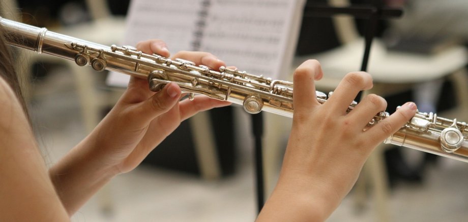 Nahaufnahme der Hände einer jungen Flötistin, die eine Querflöte spielt. Im Hintergrund sind ein Notenständer mit Notenblättern und Stühle zu sehen.