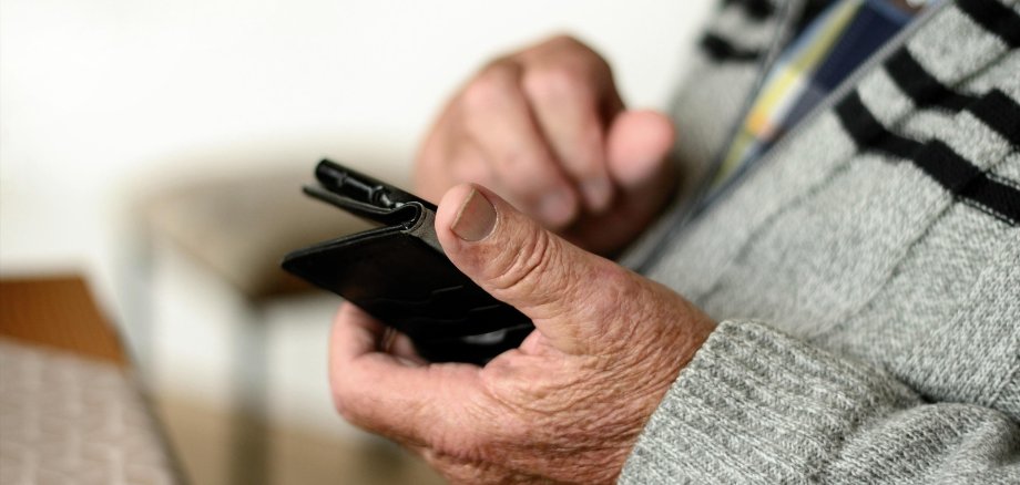 Nahaufnahme der Hände eines älteren Menschen, der ein Smartphone in einer schwarzen Schutzhülle hält und den Bildschirm mit einem Finger berührt. Im Hintergrund sind ein Tisch und eine gepolsterte Sitzbank zu erkennen.