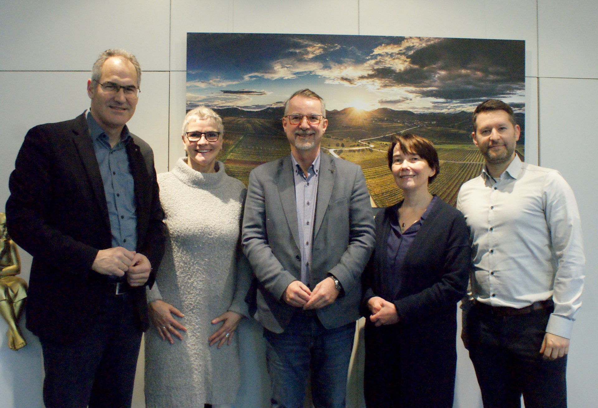 Landrat Dietmar Seefeldt, Abteilungsleiterin Michaela Endys, Ombudsmann Sven Lefkowitz, Referatsleiterin Heike Neumann und Referatsleiter Jan Oster stehen für ein Gruppenbild im Büro des Landrats nebeneinander. Hinter ihnen ist ein Bild zu sehen, dass eine Weinbergslandschaft am Rande des Pfälzerwalds bei Sonnenuntergang zeigt.