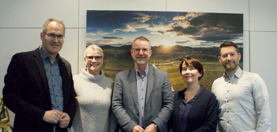 Landrat Dietmar Seefeldt, Abteilungsleiterin Michaela Endys, Ombudsmann Sven Lefkowitz, Referatsleiterin Heike Neumann und Referatsleiter Jan Oster stehen für ein Gruppenbild im Büro des Landrats nebeneinander. Hinter ihnen ist ein Bild zu sehen, dass eine Weinbergslandschaft am Rande des Pfälzerwalds bei Sonnenuntergang zeigt.
