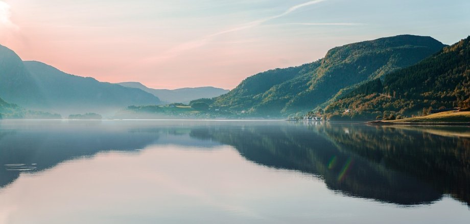 Eine ruhige See-Landschaft mit sanften Hügeln und bewaldeten Bergen im Hintergrund. Die Wasseroberfläche spiegelt den Himmel und die umliegende Natur wider. Der Himmel zeigt weiche Pastellfarben in Blau, Rosa und Gold, während sich ein feiner Nebel über das Wasser legt.