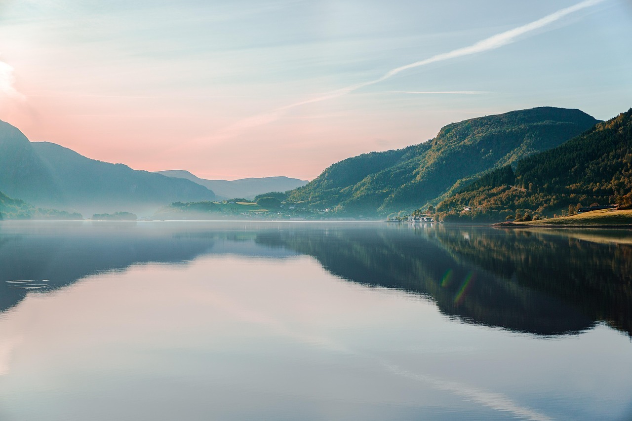 Eine ruhige See-Landschaft mit sanften Hügeln und bewaldeten Bergen im Hintergrund. Die Wasseroberfläche spiegelt den Himmel und die umliegende Natur wider. Der Himmel zeigt weiche Pastellfarben in Blau, Rosa und Gold, während sich ein feiner Nebel über das Wasser legt.