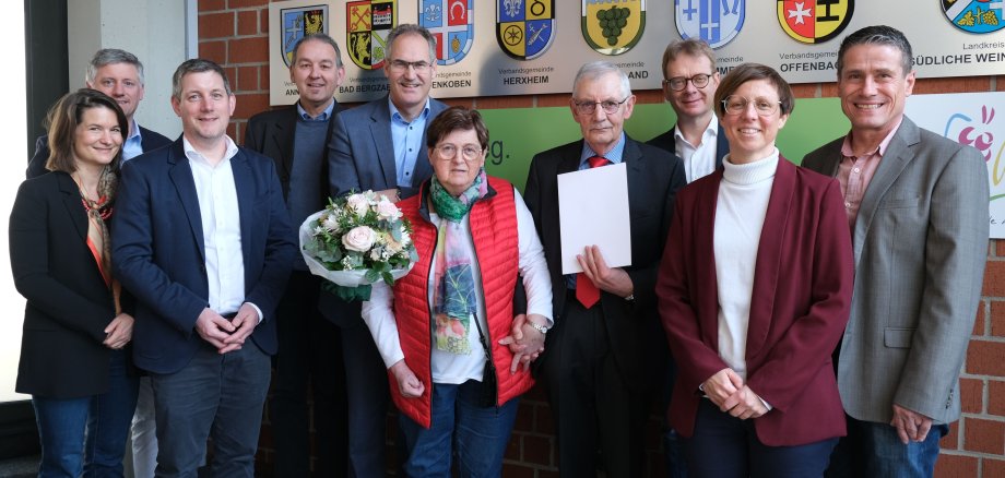 Rudolf Schwöbel (4. von rechts) im Kreis der Gratulanten und seiner Familie bei der Ehrungsfeier im Kreishaus. Foto: KV SÜW Eine Gruppe Menschen vor einer Backsteinwand mit Wappen.