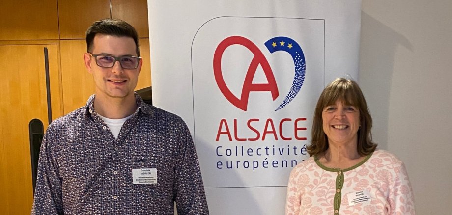 Dr. Dominik Siegler und Sabine Bader stehen vor einem Roll-Up-Banner im Hotel d'Alsace. Auf dem Banner ist das Logo der Europäischen Gebietskörperschaft Elsass (Collectivité Européenne d’Alsace) zu sehen.