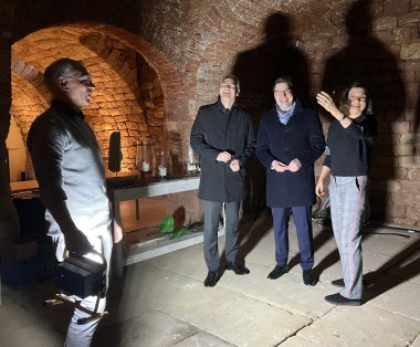 Claudia van Huet (rechts) und Bernd van Huet (vorne mit Strahler) zeigen Dietmar Seefeldt (2. von links) und Benjamin Hirsch den Weinkeller, der bald ein Konzertort sein wird. Foto: KV SÜW Eine Gruppe von vier Personen steht in einem historischen Weinkeller mit gemauerten Wänden und Gewölben. Eine Person hält einen Strahler, dessen Licht die Anwesenden sowie ihre großen Schatten an der Wand sichtbar macht. Die Gruppe schaut sich um und unterhält sich.