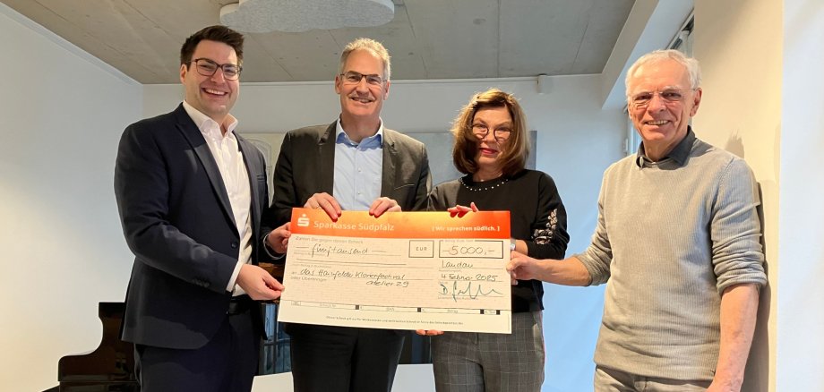 Bei der Spendenübergabe von links nach rechts: Benjamin Hirsch, Dietmar Seefeldt, Claudia van Huet und Bernd van Huet. Foto: KV SÜW Vier Personen stehen nebeneinander in einem modernen Innenraum und halten gemeinsam einen großen symbolischen Scheck über 5.000 Euro. Im Hintergrund ist ein schwarzer Flügel zu sehen. Alle blicken in die Kamera und lächeln.