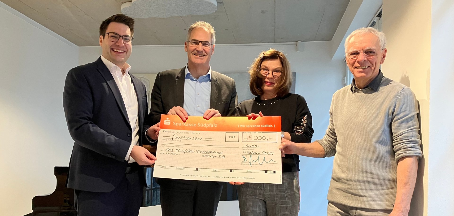 Vier Personen stehen nebeneinander in einem modernen Innenraum und halten gemeinsam einen großen symbolischen Scheck über 5.000 Euro. Im Hintergrund ist ein schwarzer Flügel zu sehen. Alle blicken in die Kamera und lächeln.