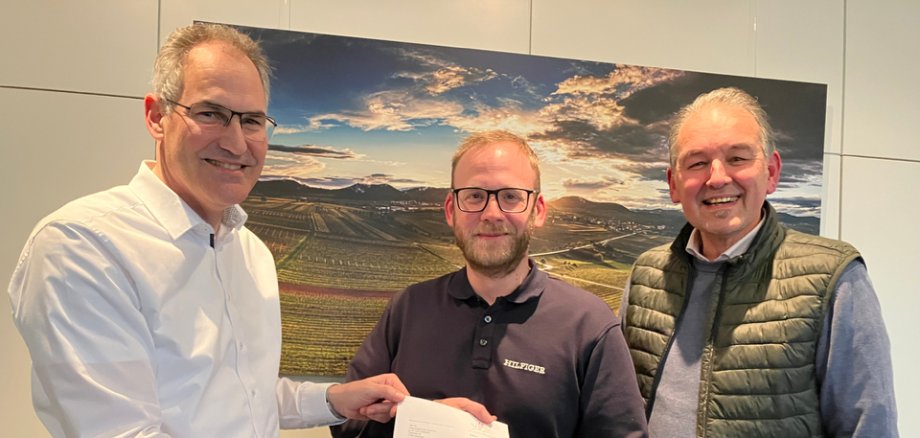 Drei Personen mit einem Bescheid vor einem Bild mit Weinbergen.