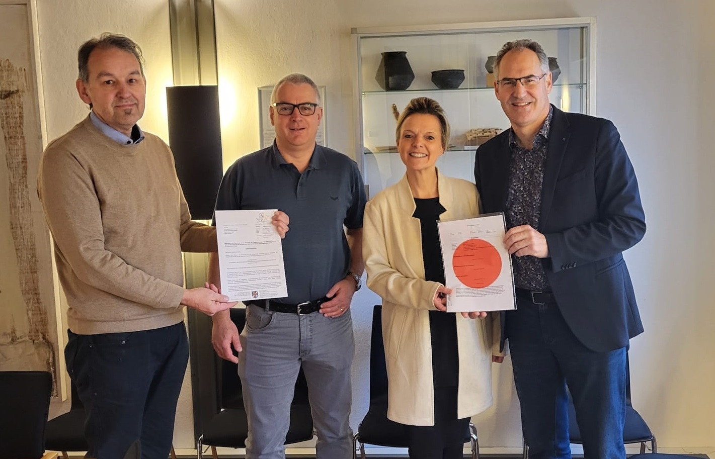 Vier Personen, zwei links halten einen Bescheid, zwei rechts ein Dokument mit rotem Punkt.