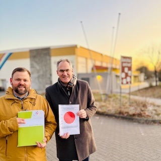 Simon Wingerter und Landrat Dietmar Seefeldt  im Porträt. Seefeldt hält den roten Punkt in der Hand, Wingerter die Unterlagen zur Baugenehmigung.