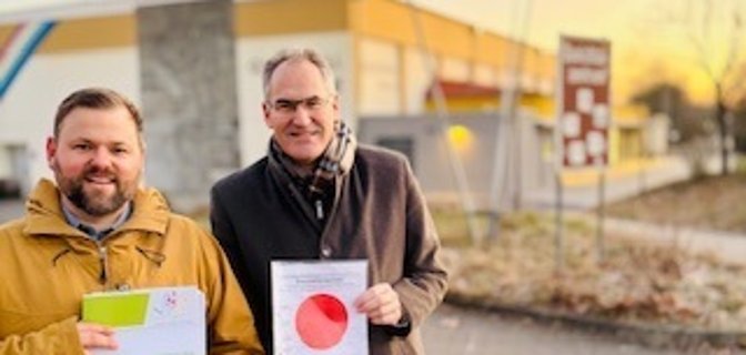 Simon Wingerter und Landrat Dietmar Seefeldt  im Porträt. Seefeldt hält den roten Punkt in der Hand, Wingerter die Unterlagen zur Baugenehmigung.
