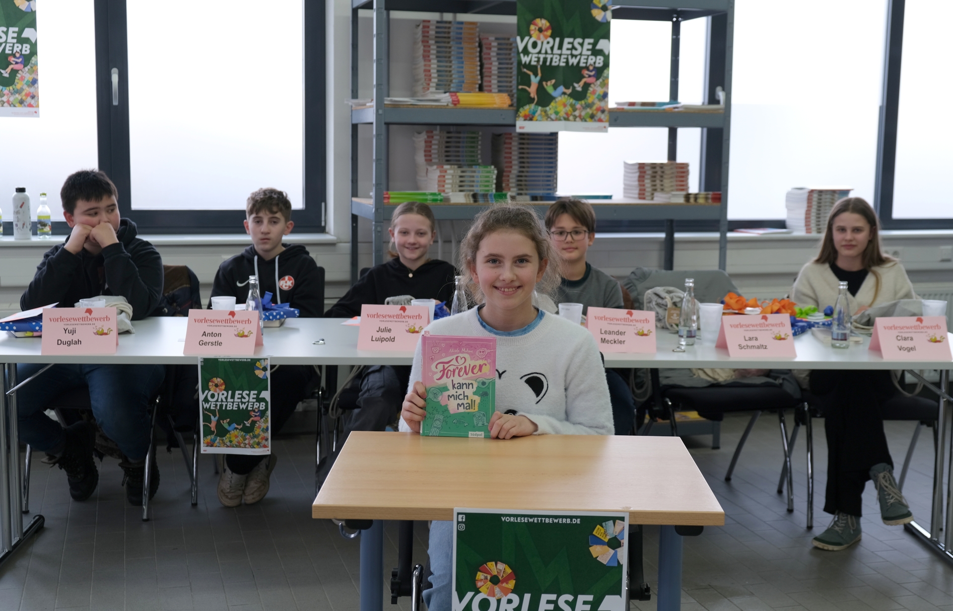 Ein Mädchen mit hellbraunen Haaren sitzt an einem Tisch und hält ein Buch mit pinkem Cover in die Kamera. Hinter ihr sitzen weitere Kinder an einem Tisch mit Namensschildern. Im Hintergrund sind Bücherregale und Plakate zum Vorlesewettbewerb zu sehen.