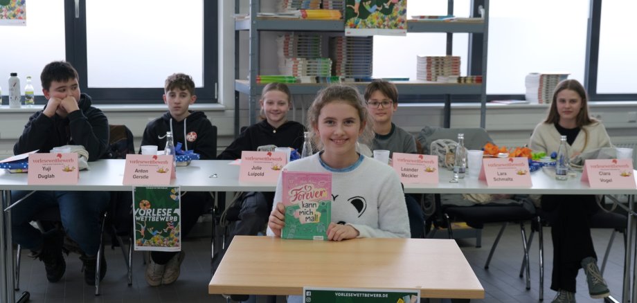 Lara Schmaltz (vorne) hat den Kreisentscheid der Südlichen Weinstraße beim diesjährigen Vorlesewettbewerb gewonnen. Foto: KV SÜW Ein Mädchen mit hellbraunen Haaren sitzt an einem Tisch und hält ein Buch mit pinkem Cover in die Kamera. Hinter ihr sitzen weitere Kinder an einem Tisch mit Namensschildern. Im Hintergrund sind Bücherregale und Plakate zum Vorlesewettbewerb zu sehen.