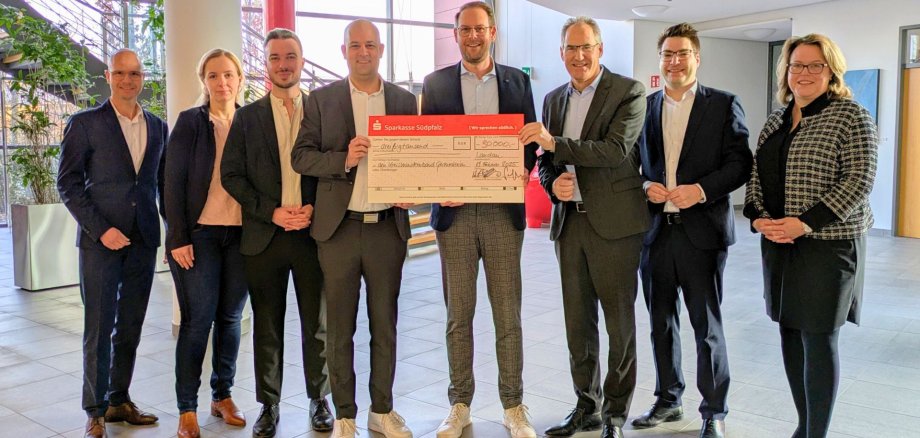 Gruppenbild: Acht Personen stehen in einem großen hellen Foyer. Die drei Personen in der MItte halten einen großen symbolischen Scheck.