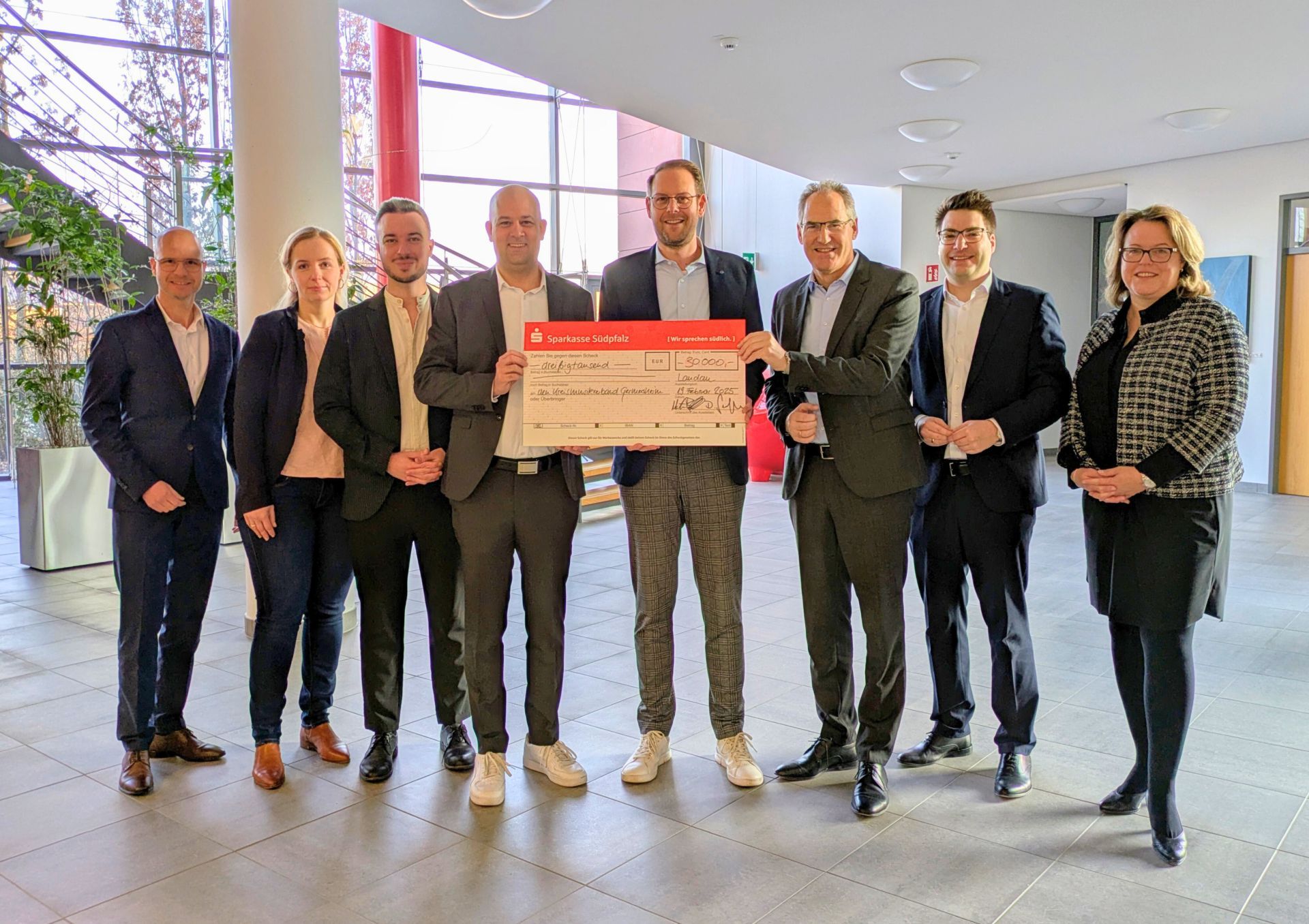 Gruppenbild: Acht Personen stehen in einem großen hellen Foyer. Die drei Personen in der MItte halten einen großen symbolischen Scheck.