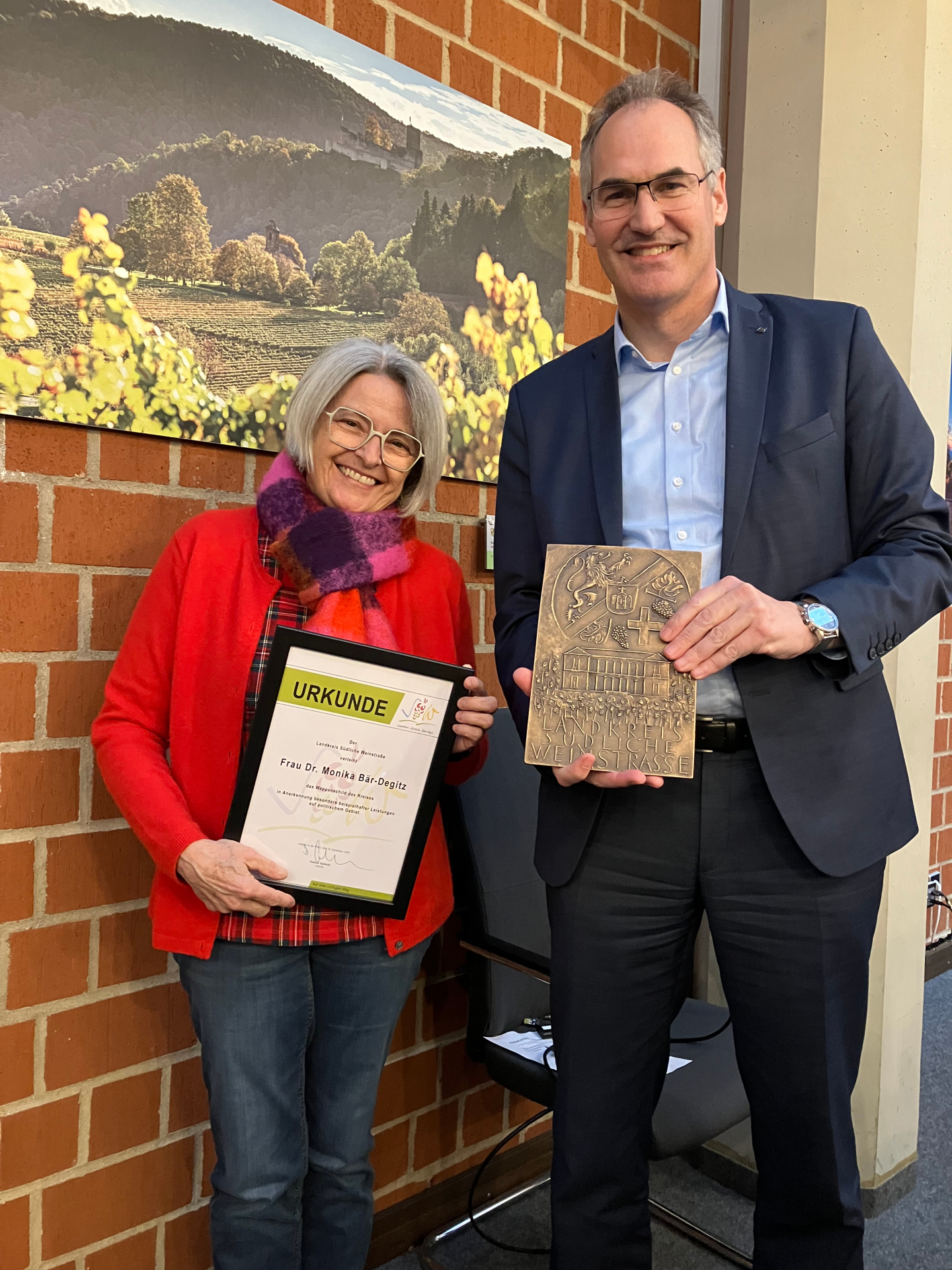 Dr. Monika Bär-Degitz und Landrat Dietmar Seefeldt  stehen im Sitzungssaal des Kreishauses vor einer Backsteinwand. Bär-Degitz hält eine Urkunde, Seefeldt den Wappenschild des Lndkreises SÜW, in Händen. Im Hintergrund hängt ein Bild an der Wand, dass eine Weinbergslandschaft zeigt.
