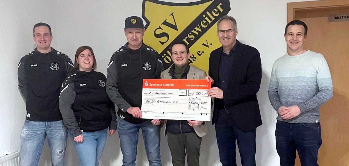 Jan Müller, Beisitzer SVV, Nina Braun, Rechnerin SVV, Andreas Müller, 1. Vorsitzender SVV, Josef Jung, Sparkasse SÜW, Landrat Dietmar Seefeldt und Bürgermeister Christian Burkhart stehen für ein Gruppenbild nebeneinander vor einer weißen Wand, auf der das Wappen des SV Völkersweilers zu sehen ist. Andreas Müller, Josef Jung und Landrat Dietmar Seefeldt halten gemeinsam den großen symbolischen Scheck.