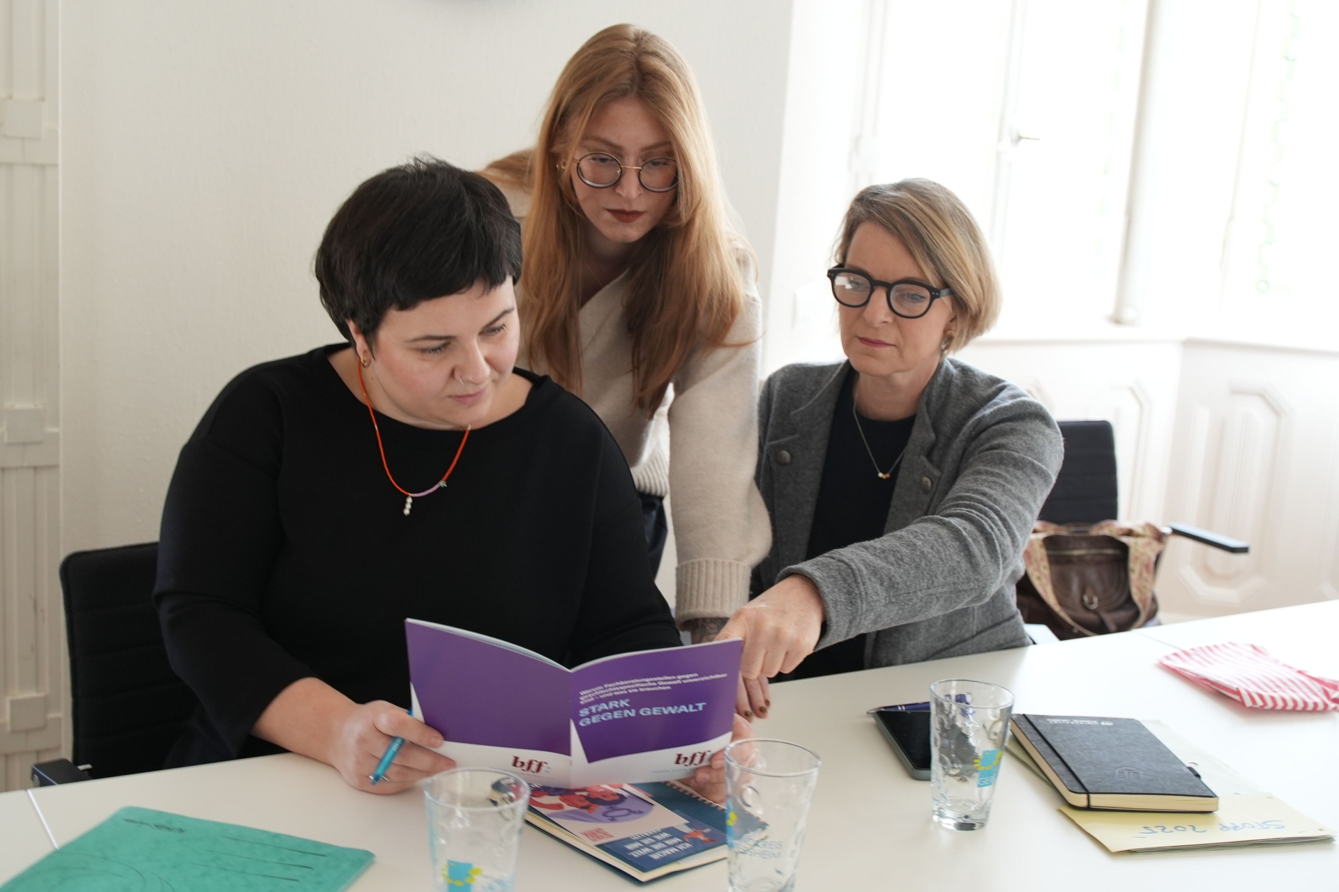 Die Gleichstellungsbeauftragten Lisa-Marie Trog, Laura Hess und Isa Stähle tauschen sich an einem Tisch aus und betrachten dabei gemeinsam eine Broschüre mit lilafarbenem Cover, auf dem die Aufschrift "Stark gegen Gewalt" zu lesen ist.
