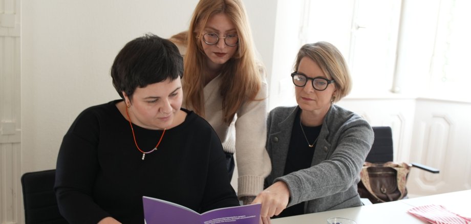 Die Gleichstellungsbeauftragten Lisa-Marie Trog, Laura Hess und Isa Stähle tauschen sich an einem Tisch aus und betrachten dabei gemeinsam eine Broschüre mit lilafarbenem Cover, auf dem die Aufschrift "Stark gegen Gewalt" zu lesen ist.