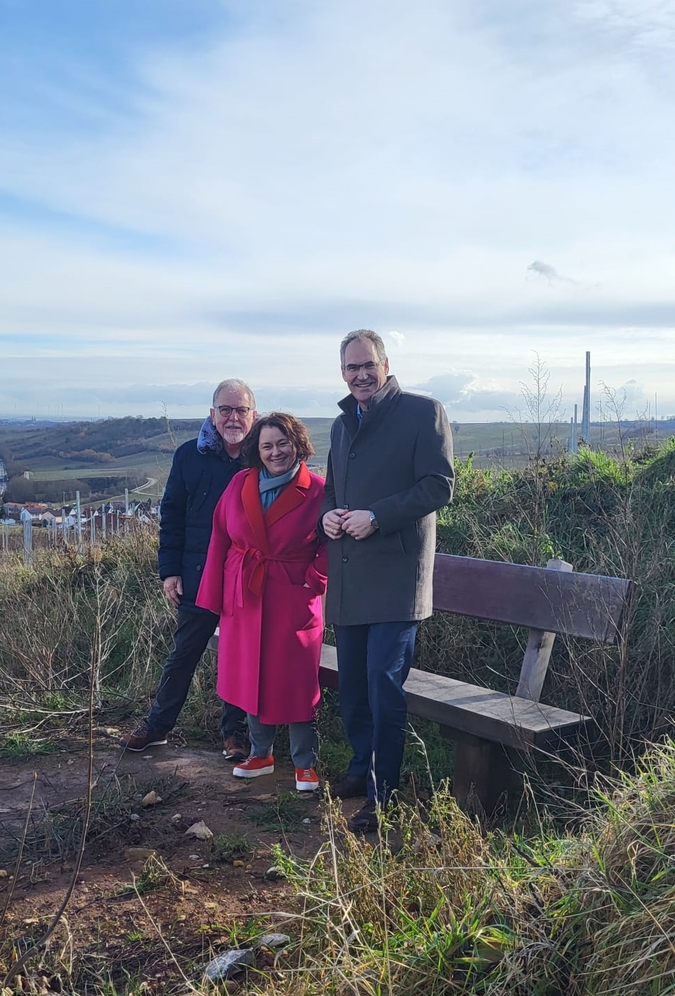 Landrat Dietmar Seefeldt, Ortsbürgermeisterin Ilse Berner und der frühere Ortsbürgermeister Bernd Flaxmeyer stehen auf einem Hügel neben einer hölzernen Bank mit Blick auf die Weinberge und das Umland von Birkweiler. Sie tragen warme Kleidung. Der Himmel ist leicht bewölkt, im Hintergrund sind sanfte Hügel, Felder und einige Windräder zu sehen.