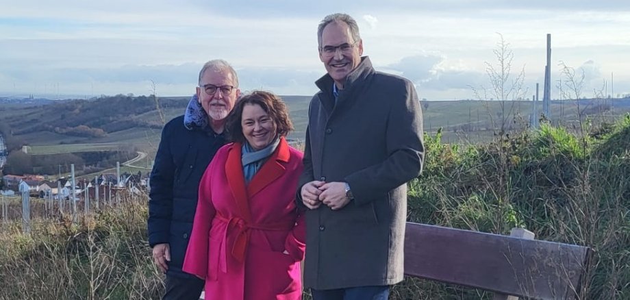 Landrat Dietmar Seefeldt, Ortsbürgermeisterin Ilse Berner und der frühere Ortsbürgermeister Bernd Flaxmeyer stehen auf einem Hügel neben einer hölzernen Bank mit Blick auf die Weinberge und das Umland von Birkweiler. Sie tragen warme Kleidung. Der Himmel ist leicht bewölkt, im Hintergrund sind sanfte Hügel, Felder und einige Windräder zu sehen.
