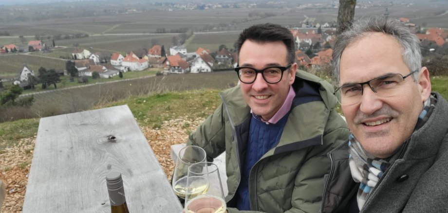 Zwei Männer an einem Holztisch stoßen mit einem Glas Weißwein an.