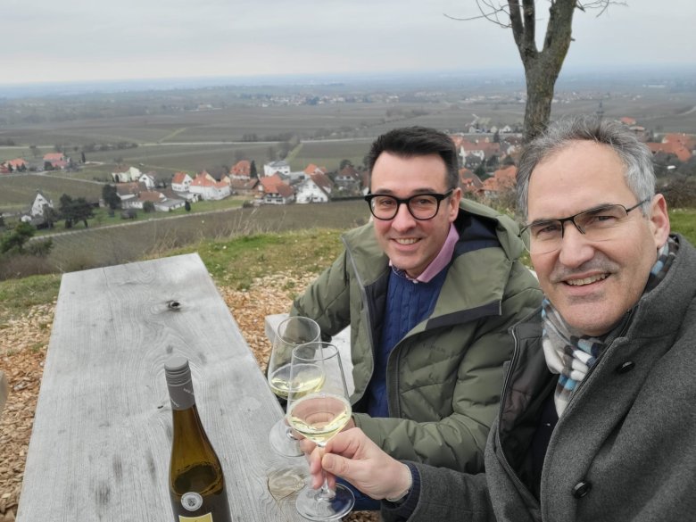Dietmar Seefeldt (rechts) und Burrweilers Ortsbürgermeister Christian Weber an der neuen Sitzgarnitur am Aussichtspunkt „Am Hüttberg“ in Burrweiler. Foto: KV SÜW Zwei Männer an einem Holztisch stoßen mit einem Glas Weißwein an.