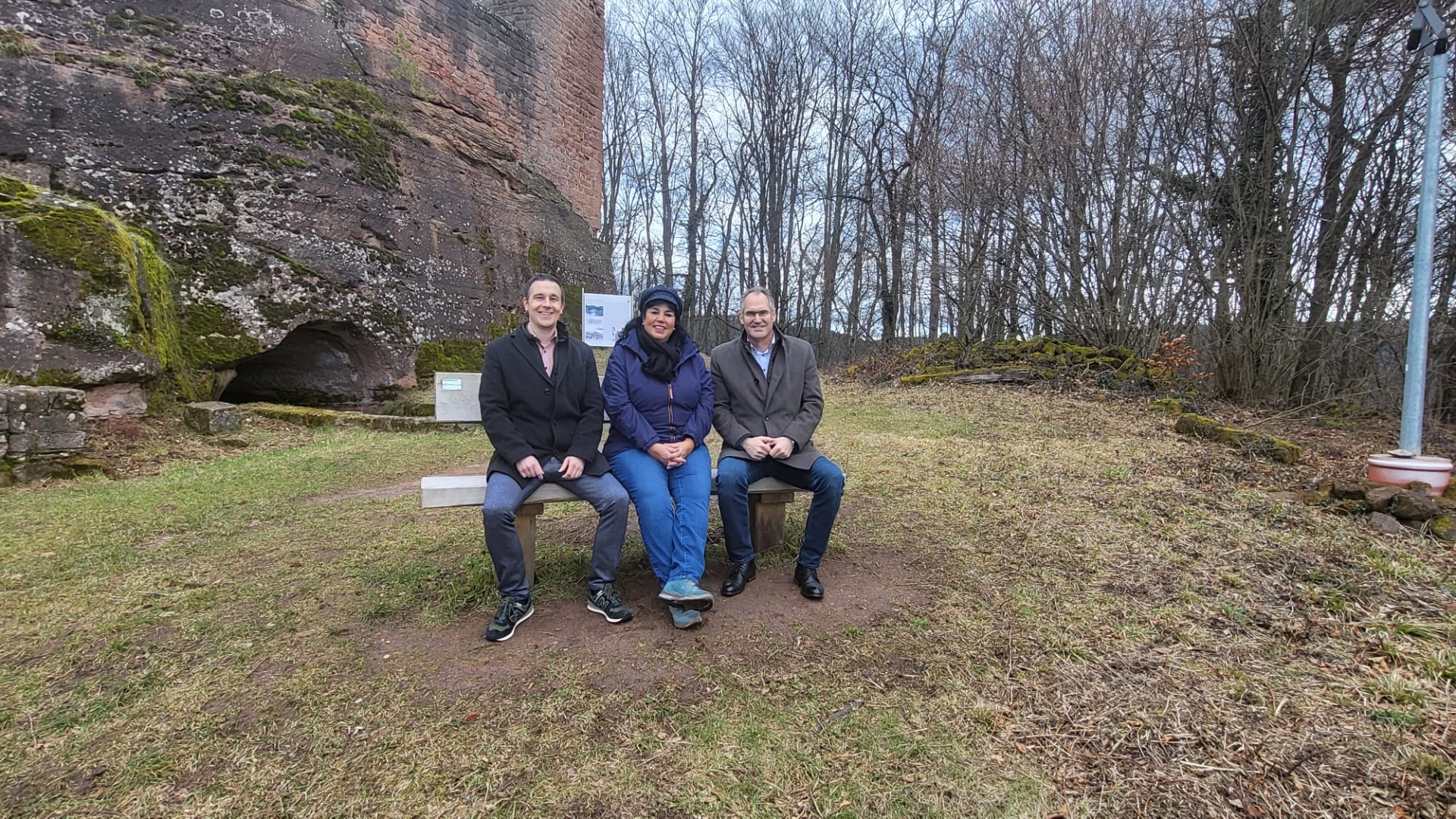 Christian Burkhart, Marina Fess und Landrat Dietmar Seefeldt sitzem nebeneinander auf einer neuen hölzernen Sitzbank unterhalb der Ramburg in Ramberg. Die Burg ist links im Hintergrund zu sehen, die ebenfalls im Hintergrund zu sehenden Bäume sind winterlich kahl.