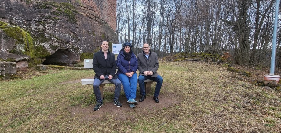 Christian Burkhart, Marina Fess und Landrat Dietmar Seefeldt sitzem nebeneinander auf einer neuen hölzernen Sitzbank unterhalb der Ramburg in Ramberg. Die Burg ist links im Hintergrund zu sehen, die ebenfalls im Hintergrund zu sehenden Bäume sind winterlich kahl.
