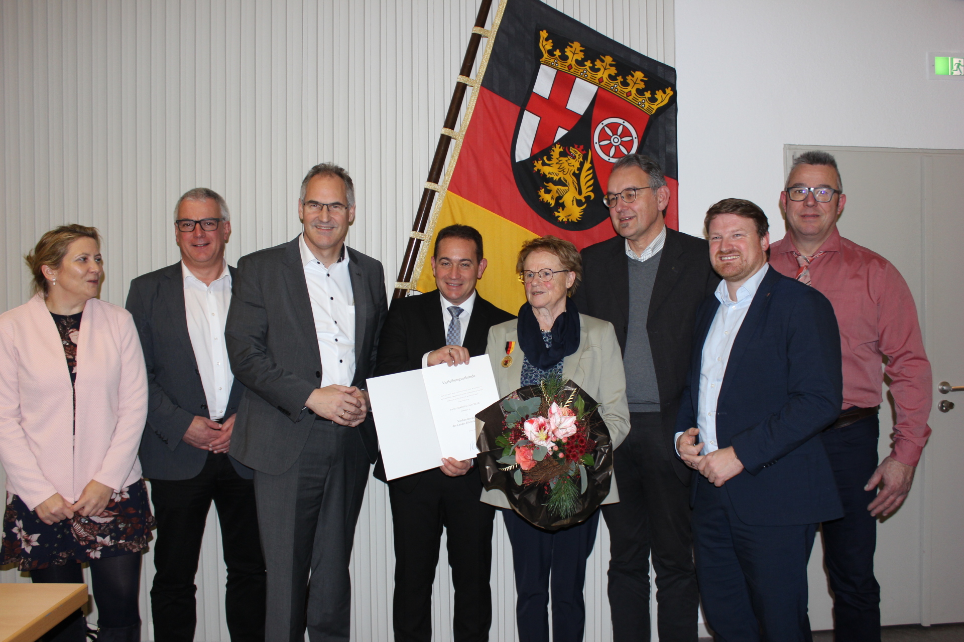 Christel Gaschler steht in einer Gruppe von Gratulanten vor der Rheinland-Pfalz-Flagge. Sie hält einen Blumenstrauß und eine Urkunde, die ihr im Rahmen einer Ehrung überreicht wurde.