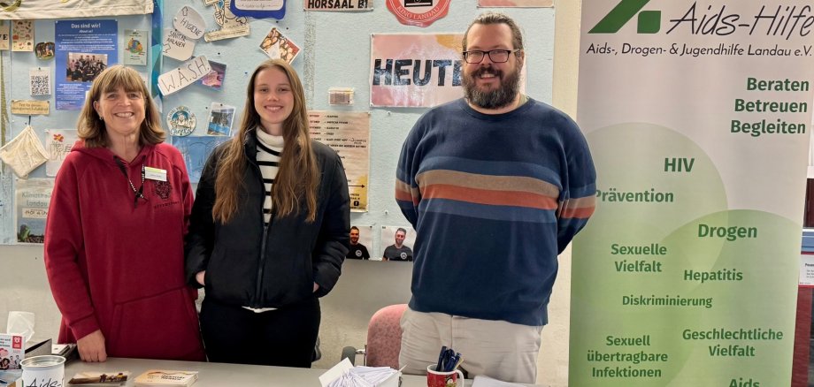 Sabine Bader vom Gesundheitsamt, Anna Staiger vom AStA und Mark Blattner von der Aids-, Drogen- und Jugendhilfe Landau e.V. stehen hinter einem Infostand mit Flyern, Broschüren und Informationsmaterialien. Rechts daneben befindet sich ein Roll-up der Aids-, Drogen- und Jugendhilfe Landau e.V., das verschiedene Themen wie HIV-Prävention, sexuelle Vielfalt und Suchtberatung auflistet. Im Hintergrund sind Poster und Plakate an einer Pinnwand zu sehen.