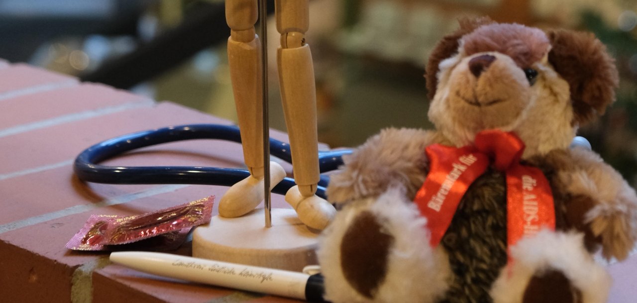 Ein hölzernes Modell eines Menschen mit einer roten Schleife in der Hand, daneben ein Teddybär mit roter Schleife. 