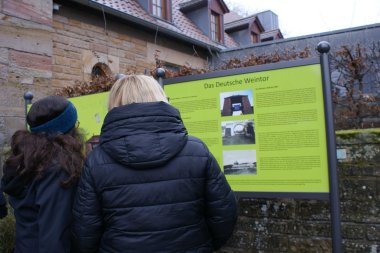 Die Tafel informiert über die Geschichte des Deutschen Weintors 1936 bis 1945. Foto: KV SÜW Nahaufnahme der Gedenktafel, die über die Geschichte des Deutschen Weintors zwischen 1936 und 1945 informiert. Zwei Personen stehen vor der Tafel, um diese zu betrachten.