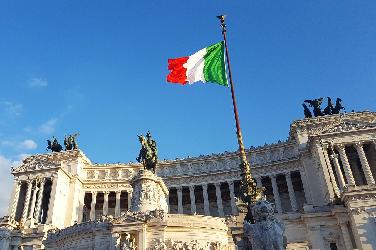 Das Monumento Nazionale a Vittorio Emanuele II in Rom, auch bekannt als Vittoriano, mit der italienischen Flagge im Vordergrund. Die weiße Marmorfassade des Nationaldenkmals ist mit Reiterstandbildern, Säulen und Quadrigen geschmückt.